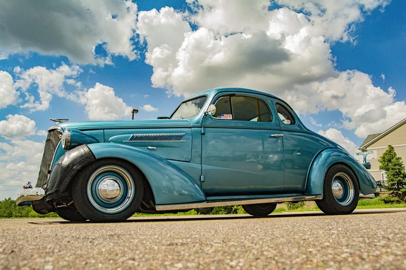 Classic teal coupe parked on a country road under a cloudy sky