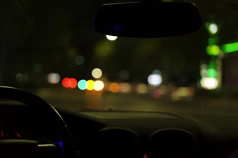 Dark windshield view at night with blurred road lights, steering wheel and dashboard