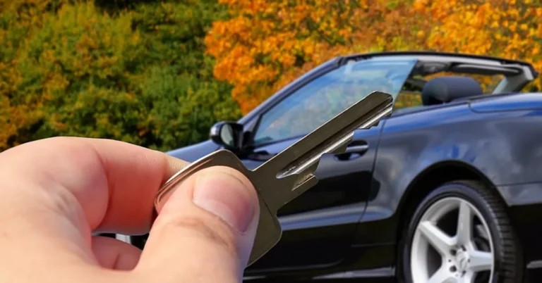 Hand holding a car key in front of a black convertible with autumn trees behind