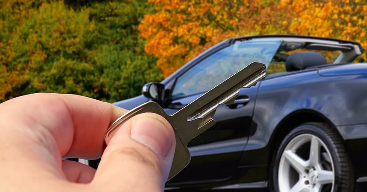 Hand holding a car key in front of a black convertible with autumn trees behind