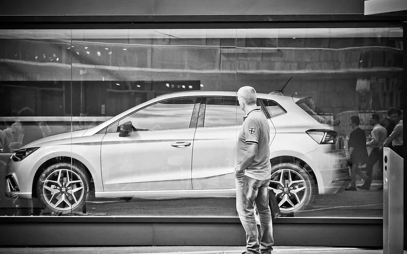 Man seen from behind looking at a white hatchback through a glass window in black and white
