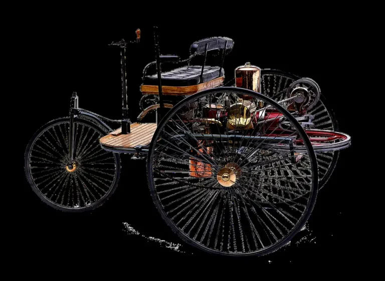 Three-wheeled early automobile with large wire-spoked wheels, a bench seat and a rear engine, shown against a black background