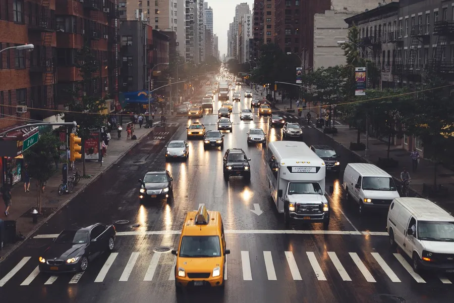 Wet city avenue at dusk with a taxi, a bus and vans stopped before a crosswalk