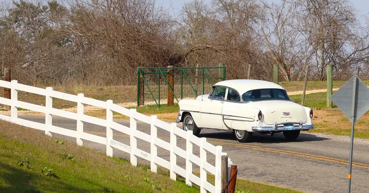 A cream colored vintage car driving along a country road beside a white fence