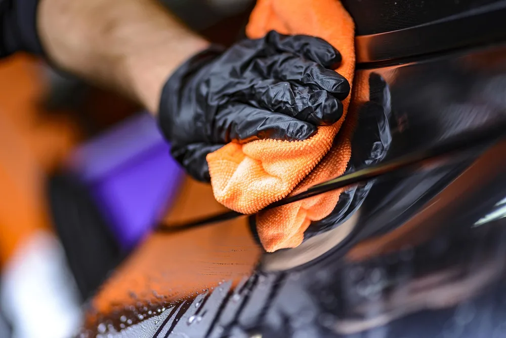 Gloved hand wiping a glossy dark car panel with an orange microfiber towel