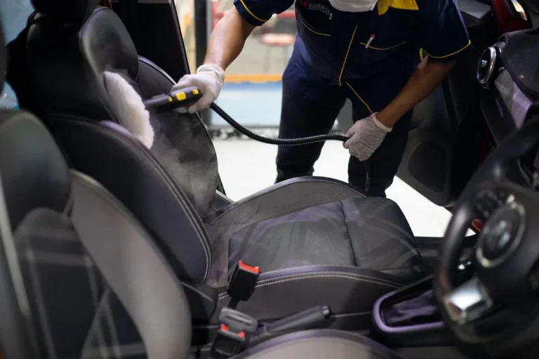 Detailer in a mask and gloves steam-cleaning a car seat with a hand steamer
