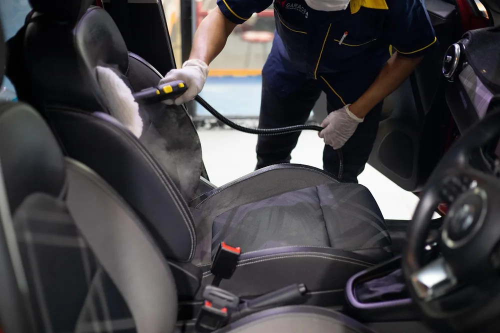 Detailer in a mask and gloves steam-cleaning a car seat with a hand steamer