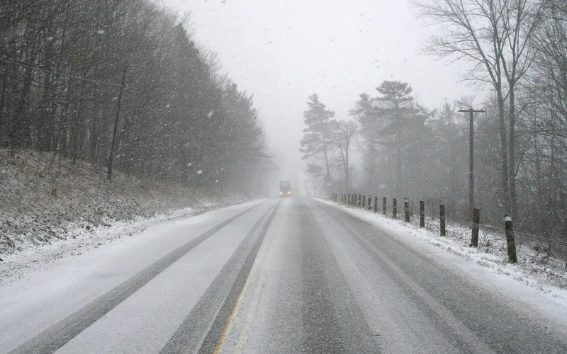 Snow-covered two-lane road in falling snow with a vehicle in the distance