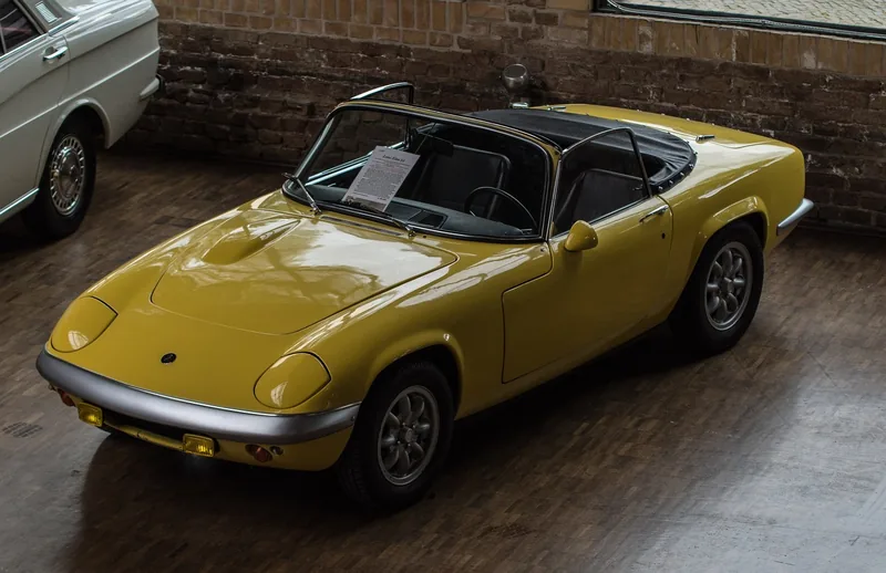 Yellow classic Lotus Elan convertible parked on a wooden floor in front of a brick wall