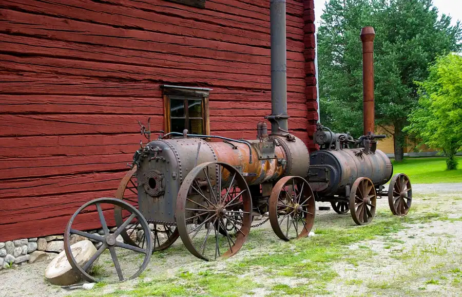 Two antique steam engines on iron wheels standing beside a red wooden barn