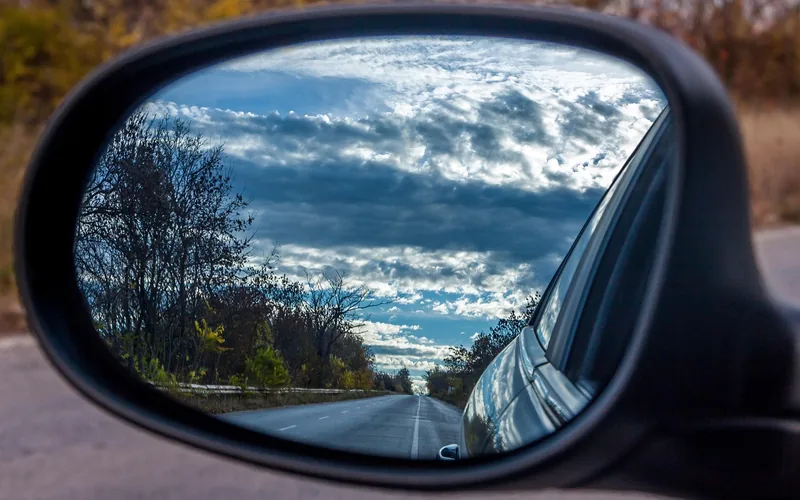 Side mirror reflecting a two-lane road and a cloudy sky