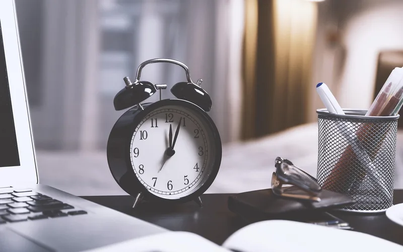 Black alarm clock on a desk next to a laptop and a pen holder