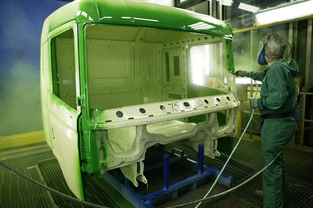 A worker in protective gear spray paints a vehicle cab inside a paint booth