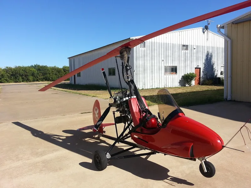 A red gyroplane with its rotor blade overhead parked on a concrete apron
