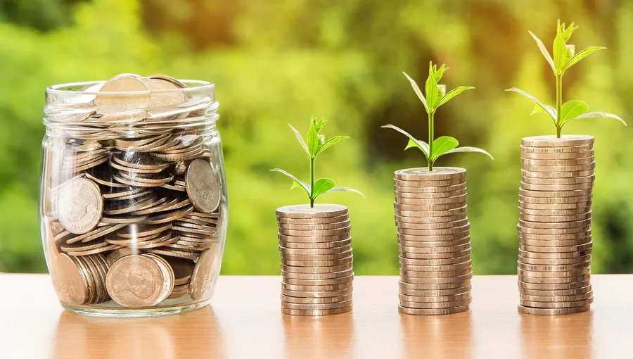 Glass jar of coins beside three stacks of coins with green seedlings growing on top