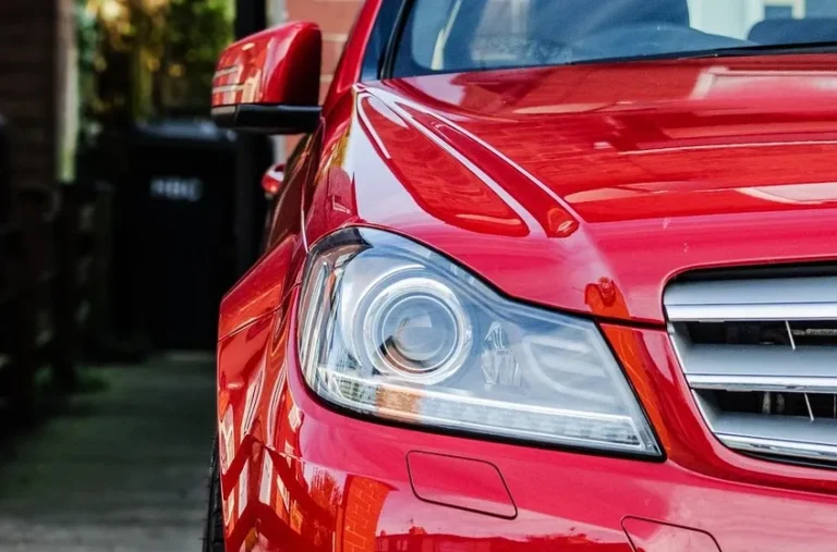 Front corner of a red car with its headlight and grille visible, parked on a gravel driveway