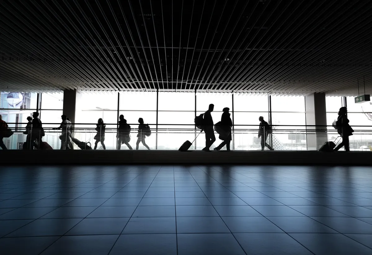 Silhouettes of travelers with luggage walking through an airport terminal with large windows