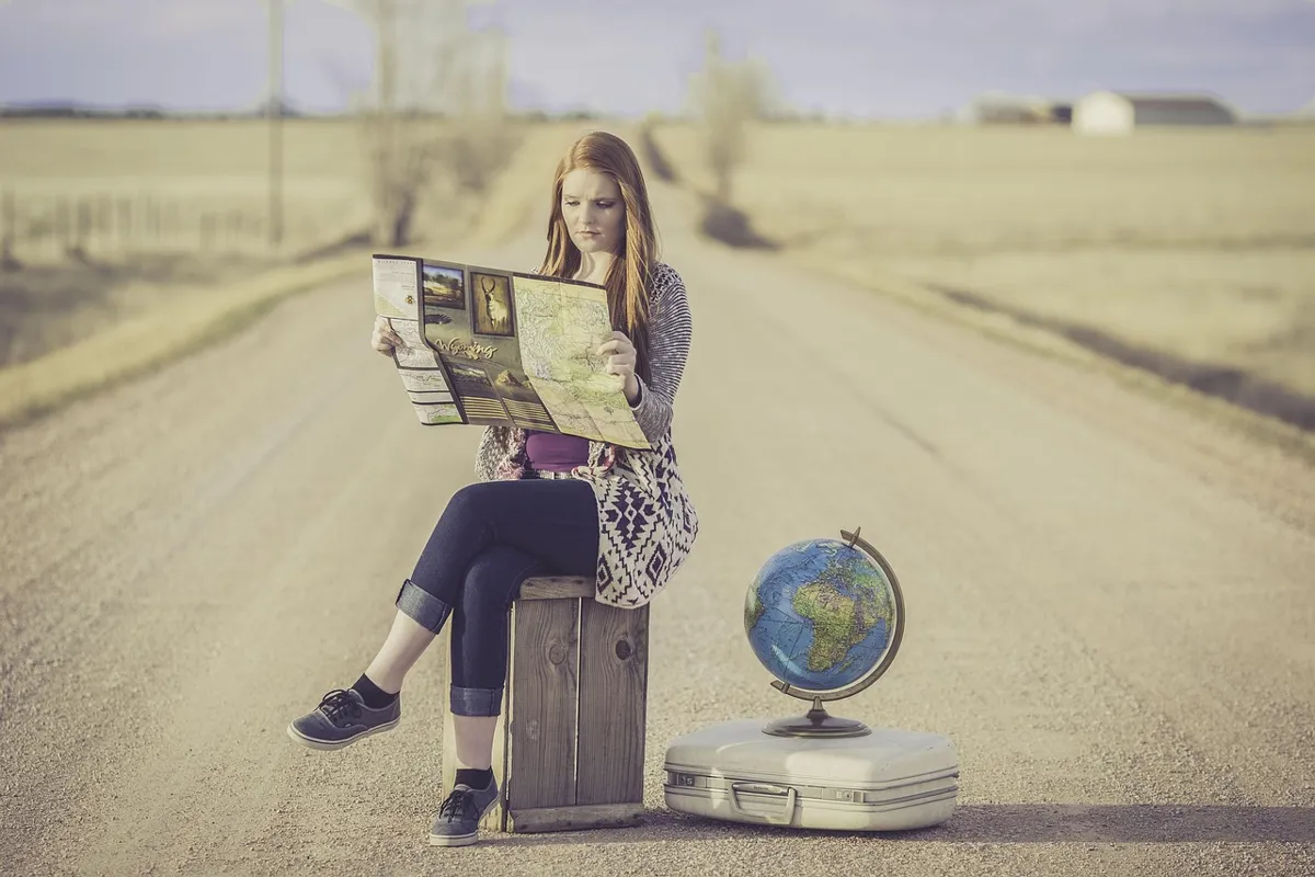 Traveler sitting on a wooden crate on a rural road reading a paper map next to a globe