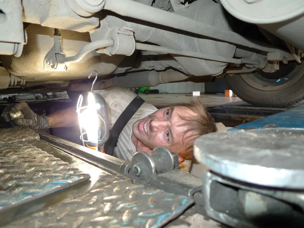 A mechanic with a work light looking at the underside of a vehicle in a shop