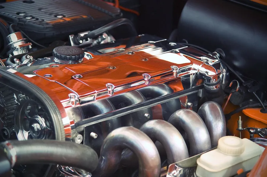 Open engine bay of a car showing a chrome valve cover, exhaust manifold and rubber hoses