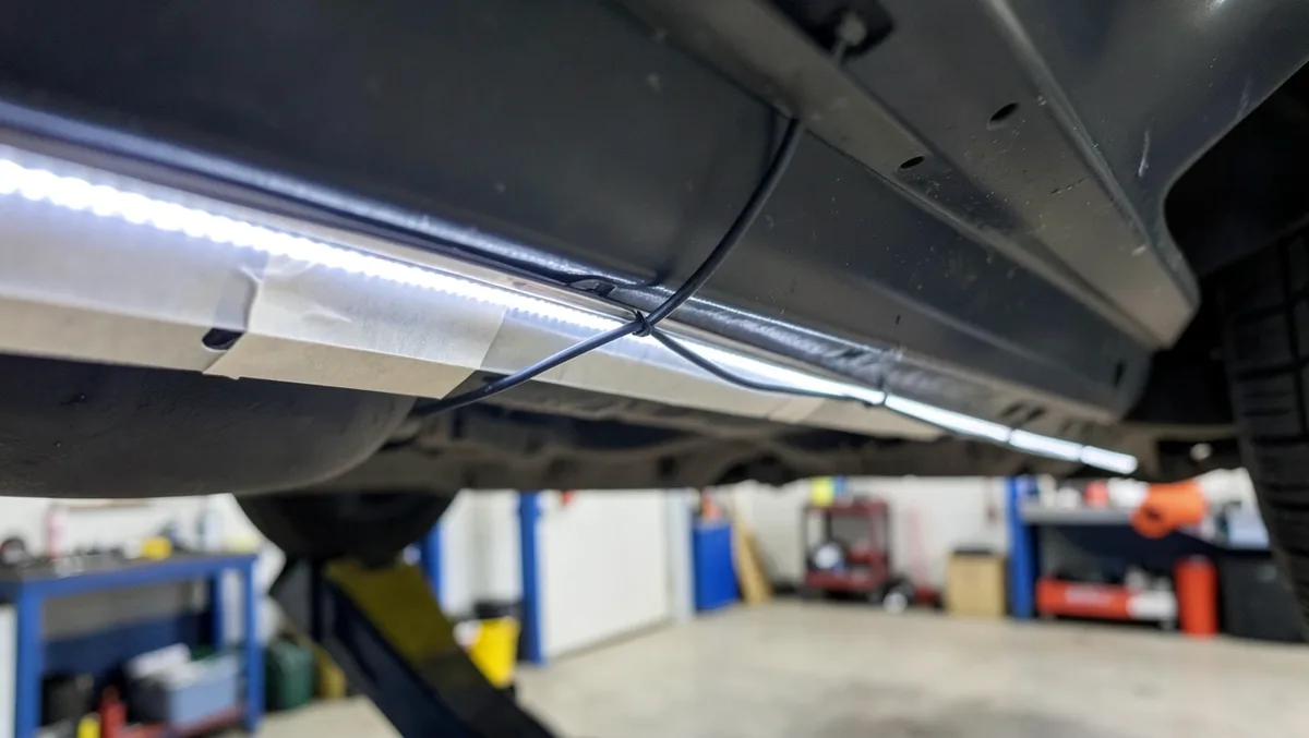 Close-up of an LED underglow light strip taped along a car rocker panel with wiring zip-tied to the frame