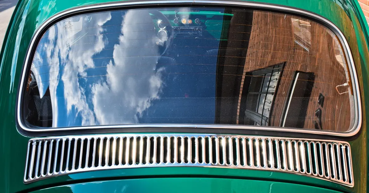 Rear window of a green vintage car reflecting clouds and a brick building