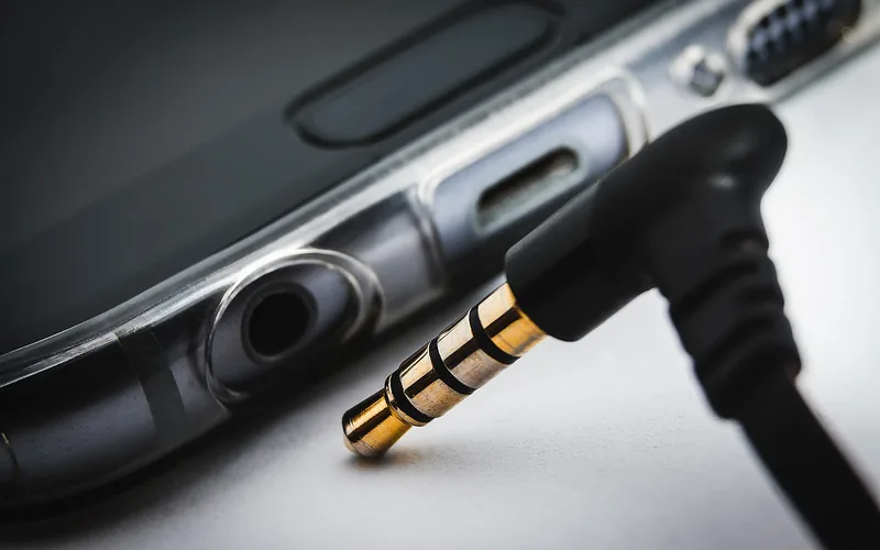 A 3.5 mm audio plug with gold rings next to a phone with a headphone jack and charging port