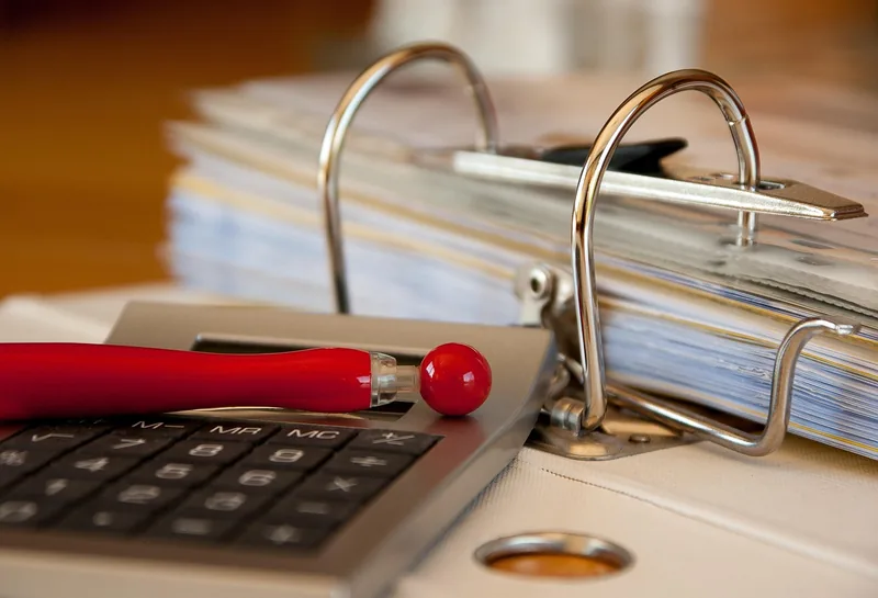 Binder of paperwork beside a calculator and a red pen