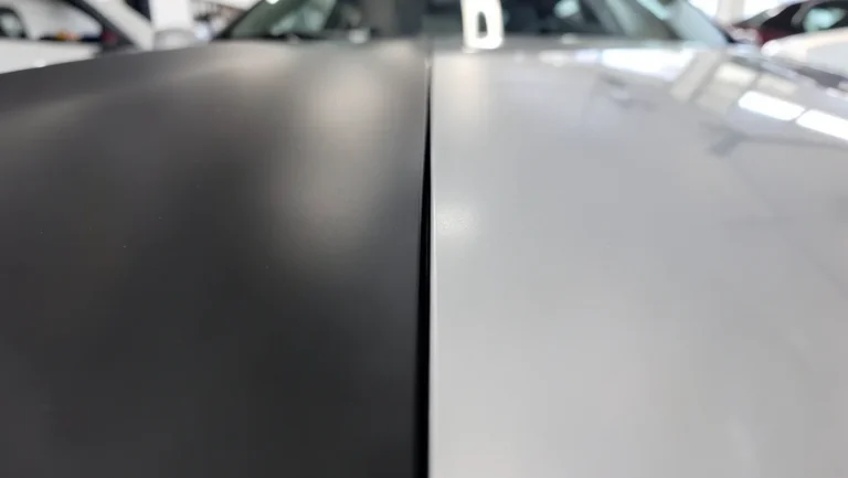 Close-up comparison of matte black vinyl wrap film next to glossy factory paint on a car hood, showing the seam where the wrap ends
