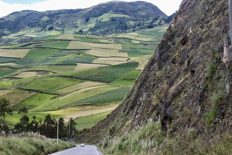 A car on a road running beside a steep mountain slope above farm fields