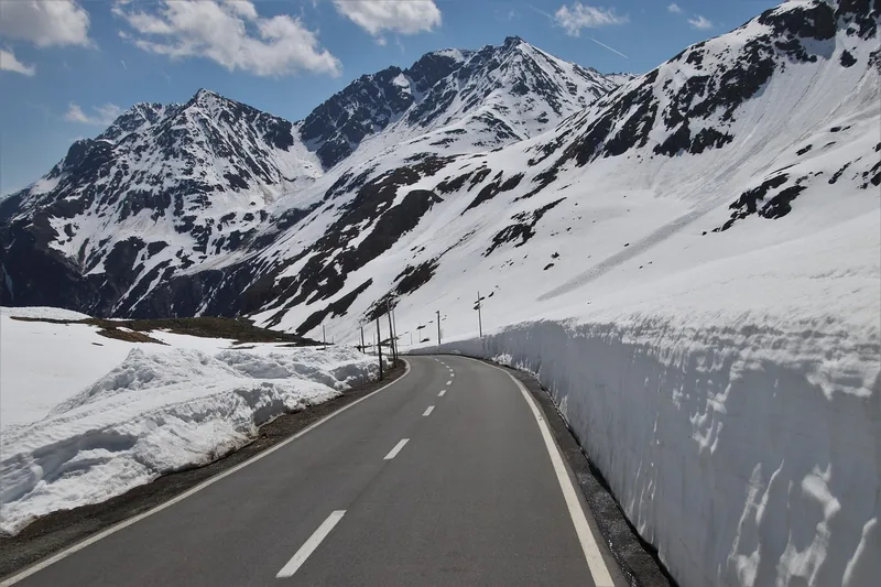 Cleared mountain road between tall snow banks with snowy peaks behind