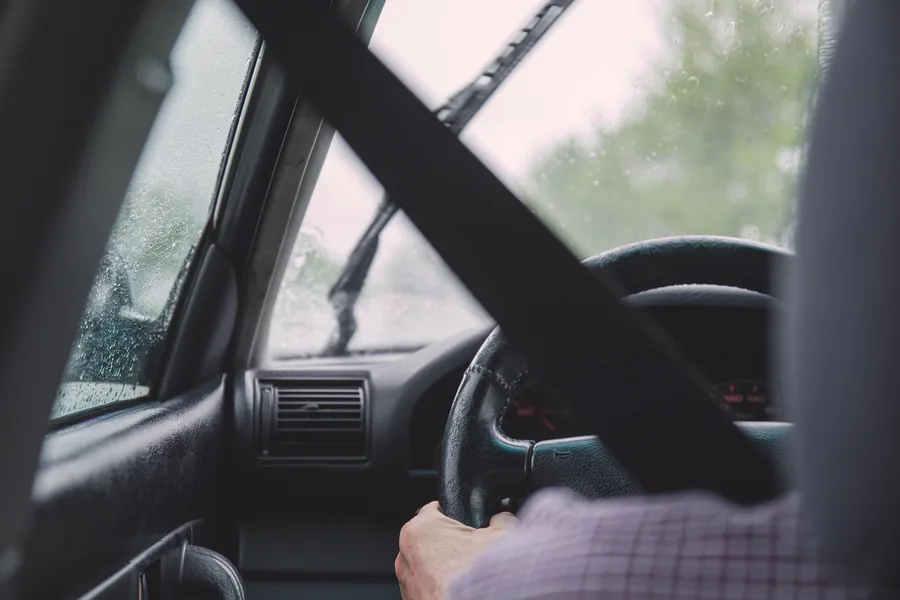 Driver with a hand on the steering wheel in the rain, seat belt across the frame