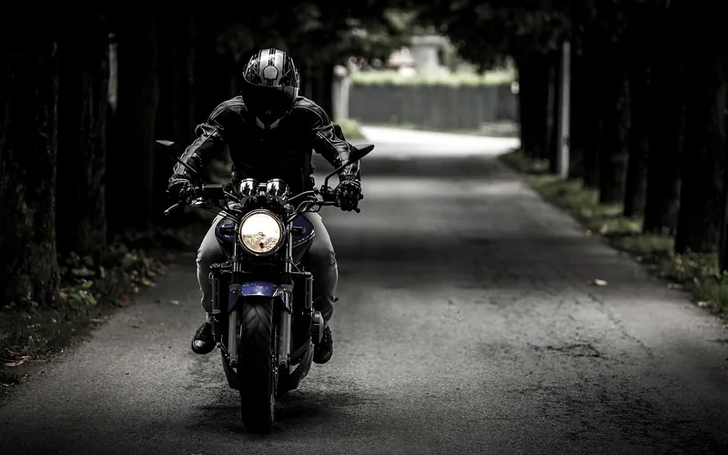 Motorcyclist in a helmet and leather jacket riding on a tree-lined road with the headlight on
