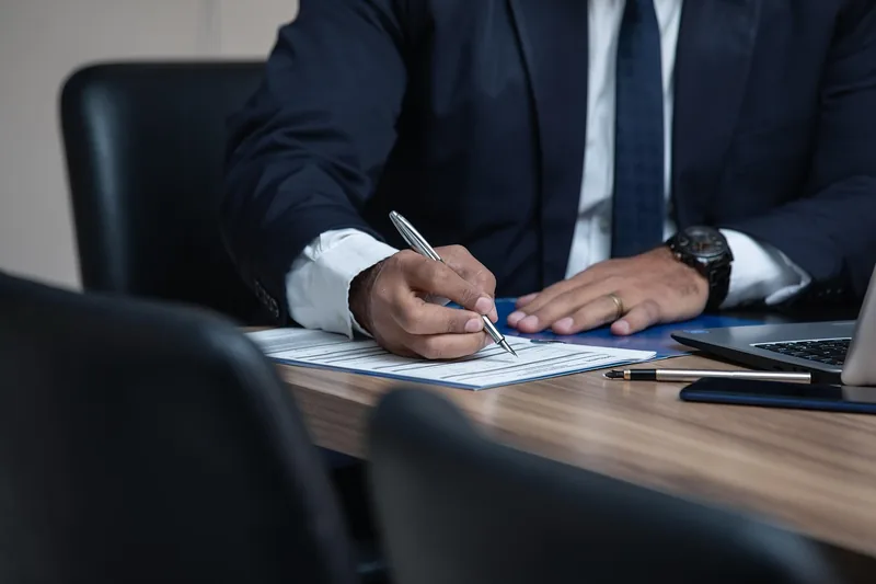 Person in a suit signing a form at a desk with a laptop and a pen nearby