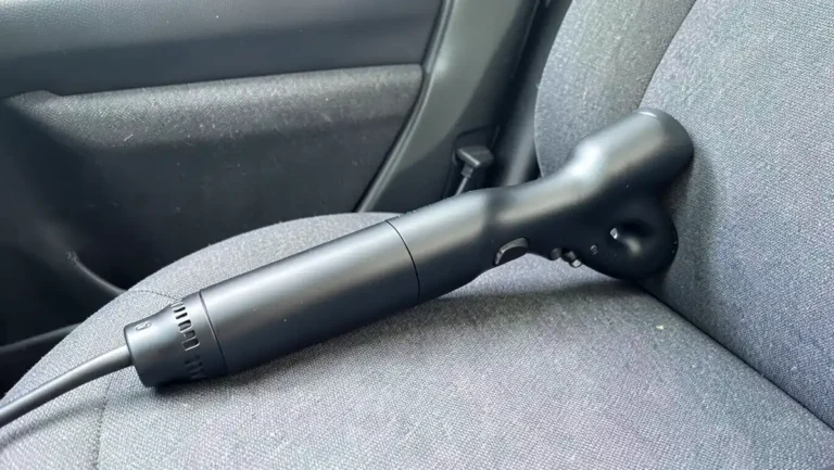 Handheld 12V vacuum cleaner resting on a gray car seat with its cord running to the dashboard socket