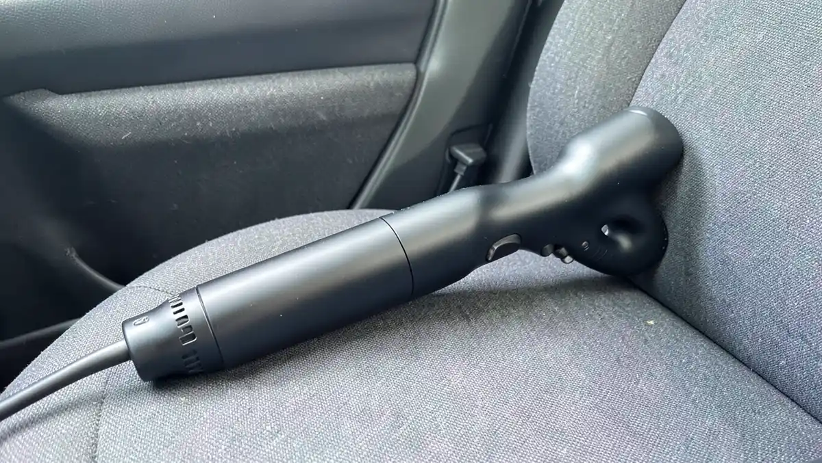Handheld 12V vacuum cleaner resting on a gray car seat with its cord running to the dashboard socket