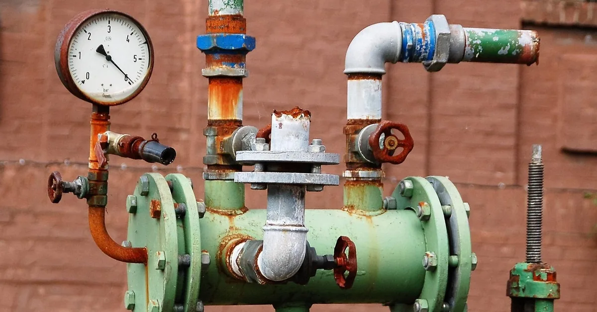 Round pressure gauge mounted above red hand valves on a rusty green pipe assembly