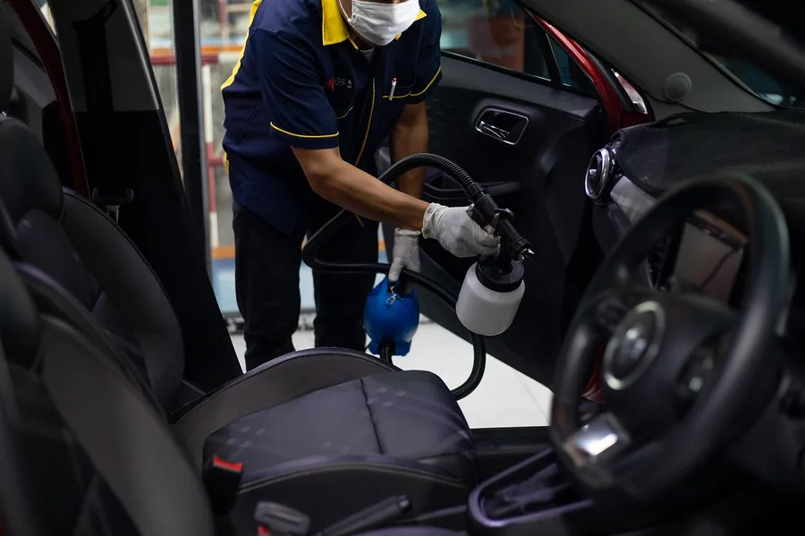 Worker in gloves and mask treating a car seat with a spray tool