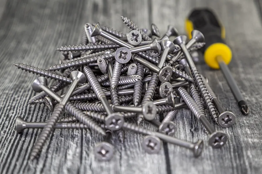 Pile of Phillips-head screws on a gray wooden surface with a screwdriver beside them