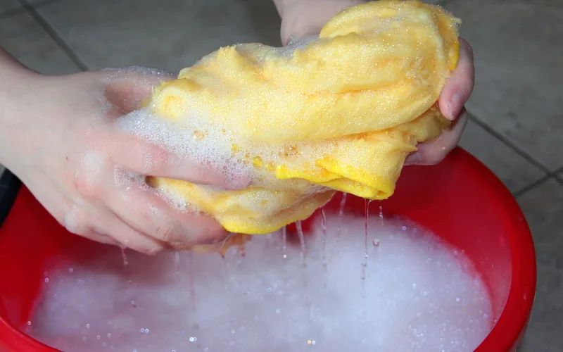 Hands wringing a soapy yellow sponge over a red wash bucket