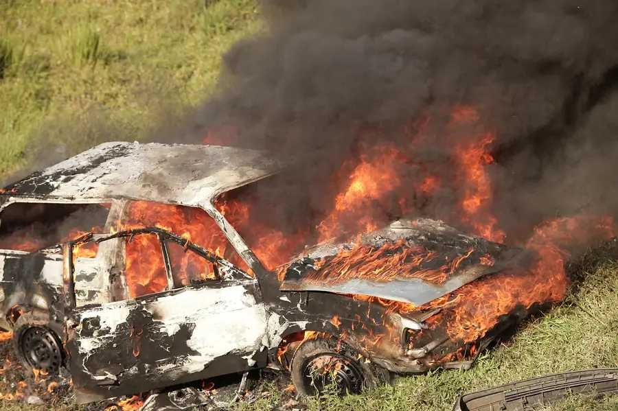 Car on fire in a grassy field with flames and black smoke coming from the hood and windows