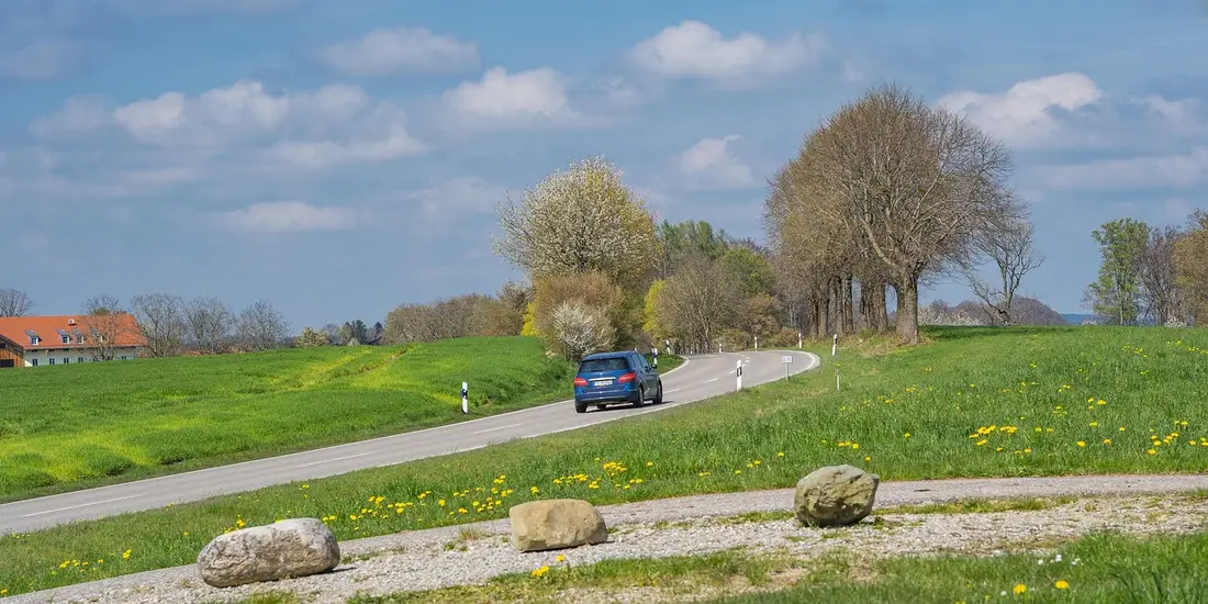 A blue car driving along a curving country road past green fields