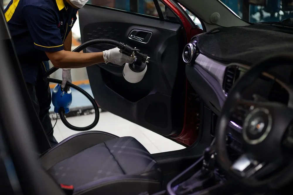 A worker in a uniform and gloves uses a spray tool on a car door panel during an interior cleaning