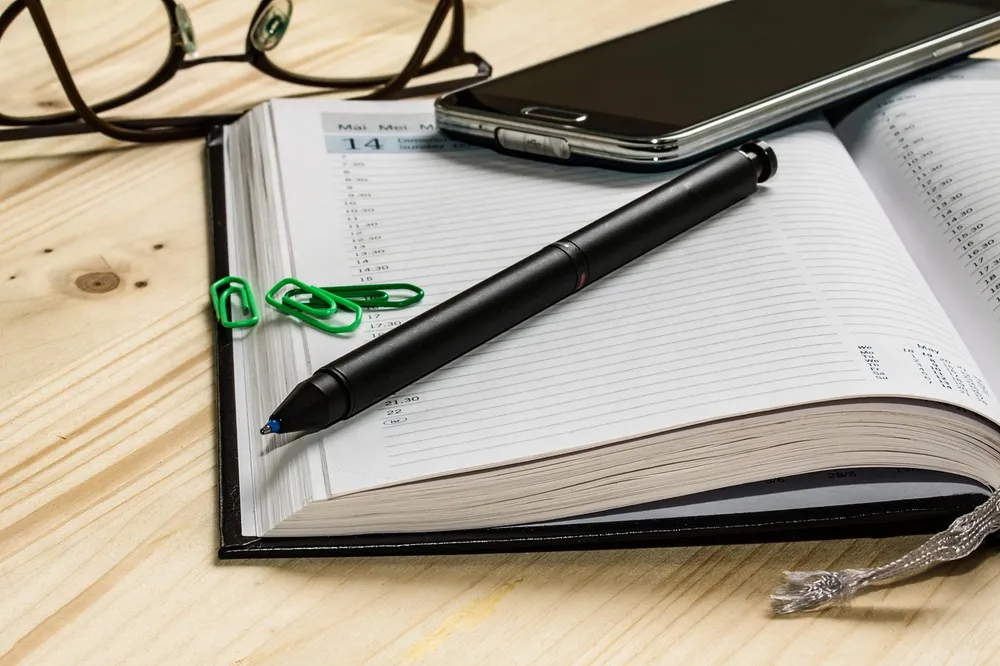 Open paper planner with a pen, paper clips and a smartphone on a wooden desk