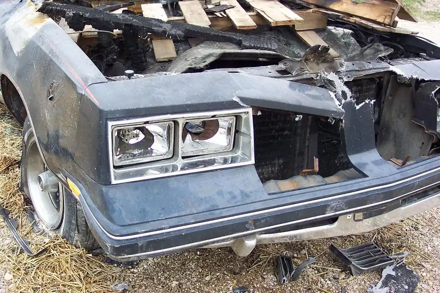 Front end of a dark car with a burned engine bay, a broken headlight unit and a crumpled hood
