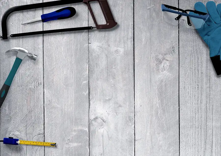 Safety glasses and a blue work glove beside a hacksaw, hammer, screwdriver and tape measure on wooden boards