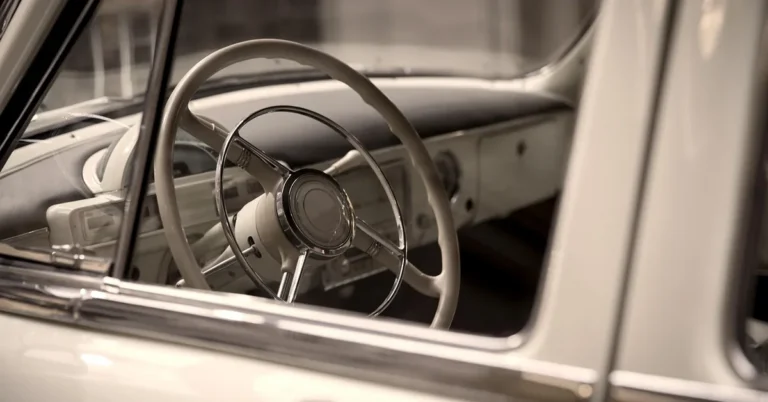 The steering wheel and dashboard of a vintage cream-colored car seen through the side window