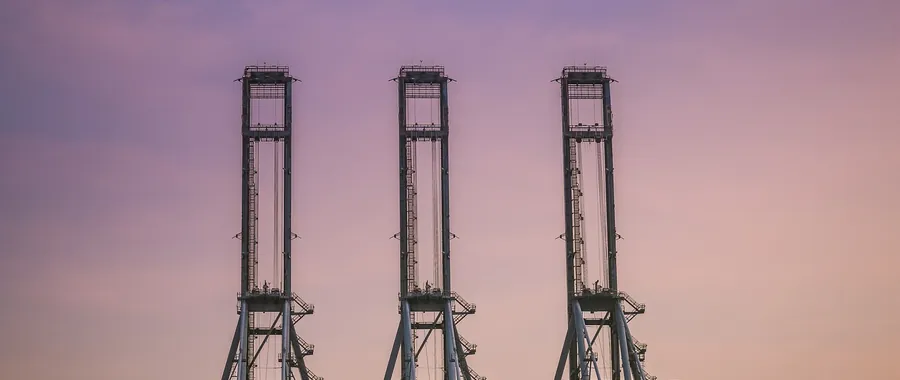 Three container gantry cranes standing against a dusk sky at a cargo port