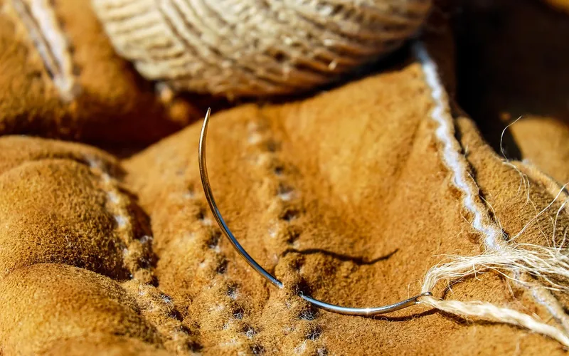 Curved upholstery needle with thread resting on a stitched suede leather piece