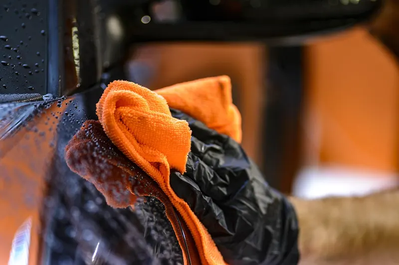 Gloved hand wiping a wet car surface with an orange microfiber cloth